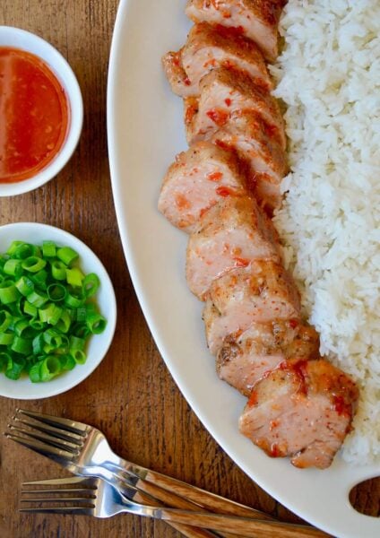 Scallions and Thai sweet chili sauce in small white bowls next to two forks and white serving tray with Sweet and Sour Roast Pork Tenderloin with white rice.