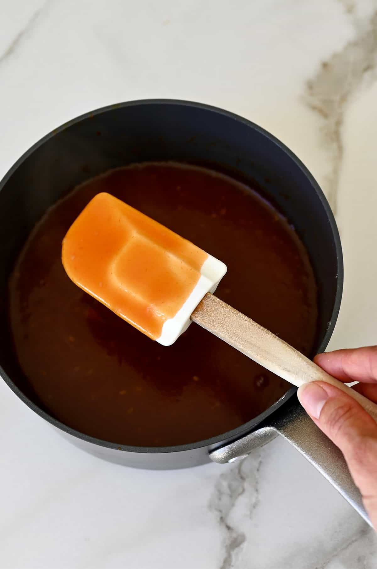 A hand holds a spatula covered in sweet and sour sauce over a saucepan containing more sauce.