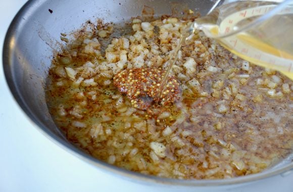 Large sauté pan with diced onions and whole grain mustard with apple cider vinegar being poured from glass measuring cup.