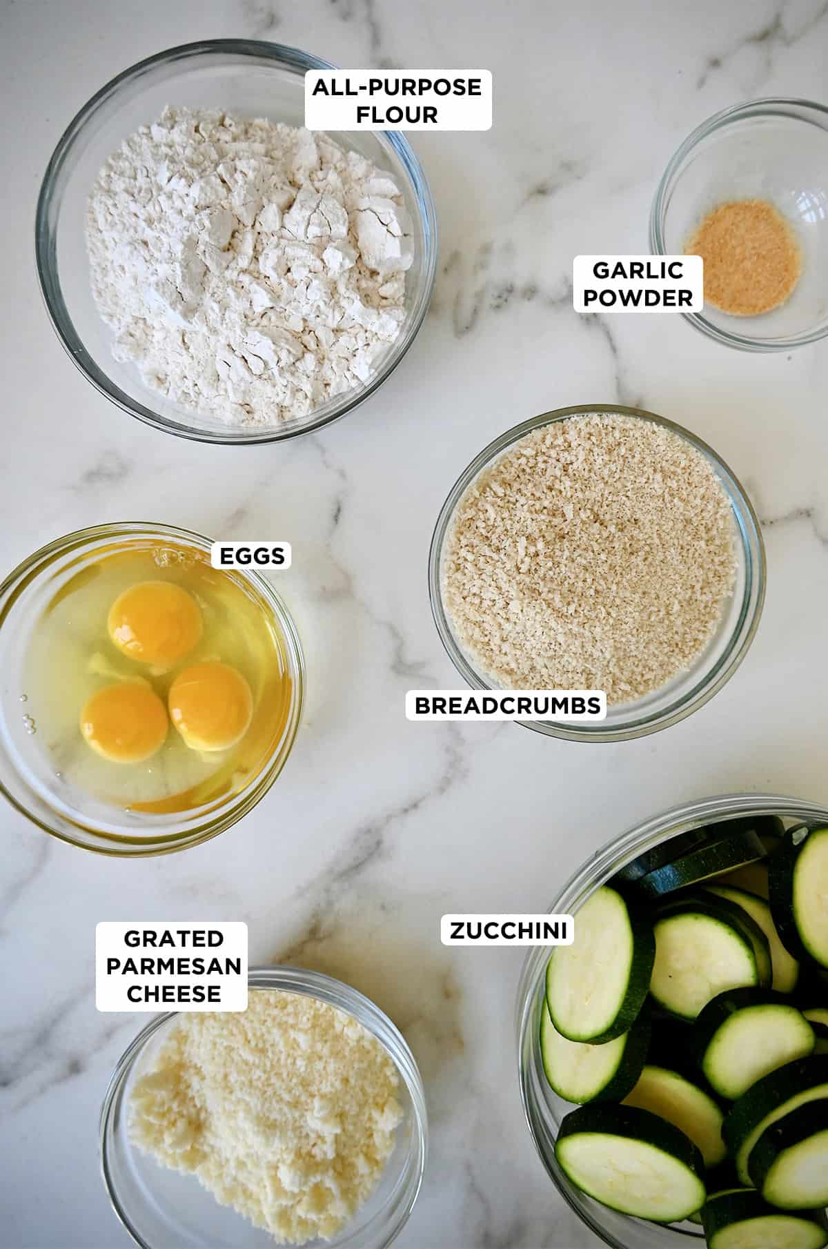 Ingredients to make zucchini chips in glass bowls, including sliced zucchini, grated Parmesan cheese, eggs, breadcrumbs, flour and garlic powder.