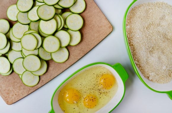 Cutting board with zucchini chips, small white and green bowl with three eggs, and a medium white and green bowl with breadcrumbs.