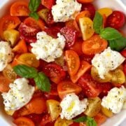 A white bowl containing red, orange and yellow halved tomatoes, burrata and basil