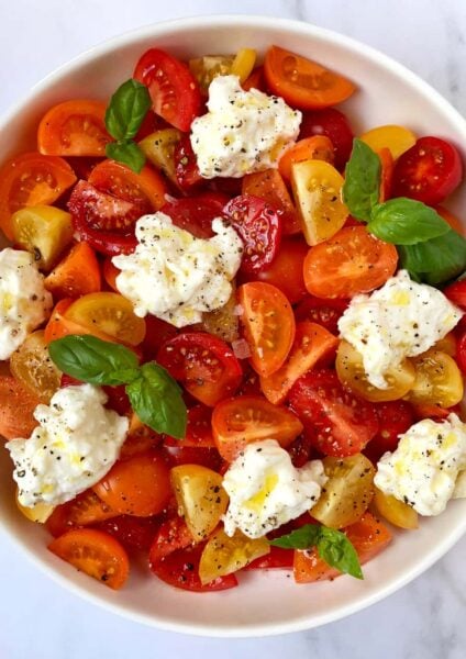 A white bowl containing red, orange and yellow halved tomatoes, burrata and basil