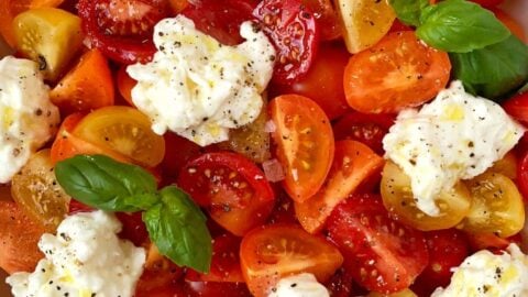 A white bowl containing red, orange and yellow halved tomatoes, burrata and basil