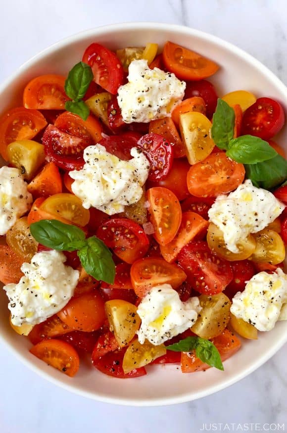 A white bowl containing red, orange and yellow halved tomatoes, burrata and basil