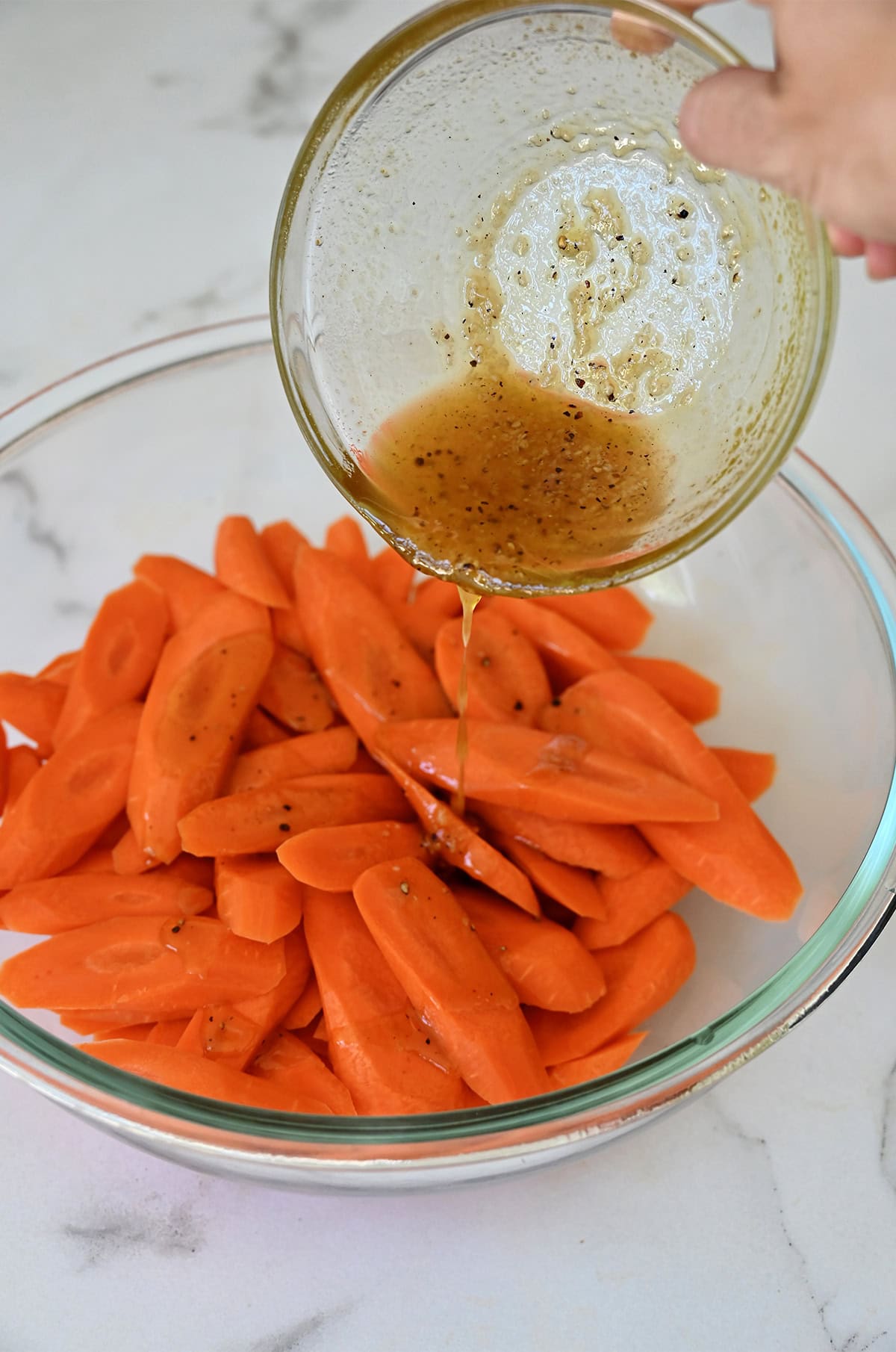 A honey-olive oil mixture with salt and pepper being poured from a small bowl over sliced carrots in a larger bowl.