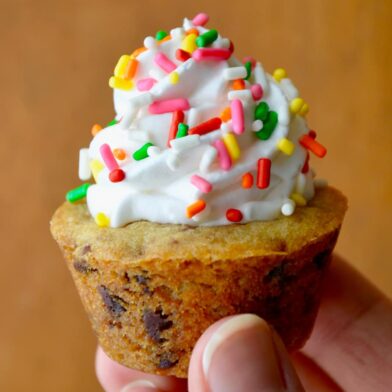A close-up shot of a hand holding a Chocolate Chip Cookie Cup topped with vanilla frosting and sprinkles