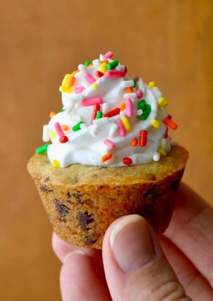A close-up shot of a hand holding a Chocolate Chip Cookie Cup topped with vanilla frosting and sprinkles