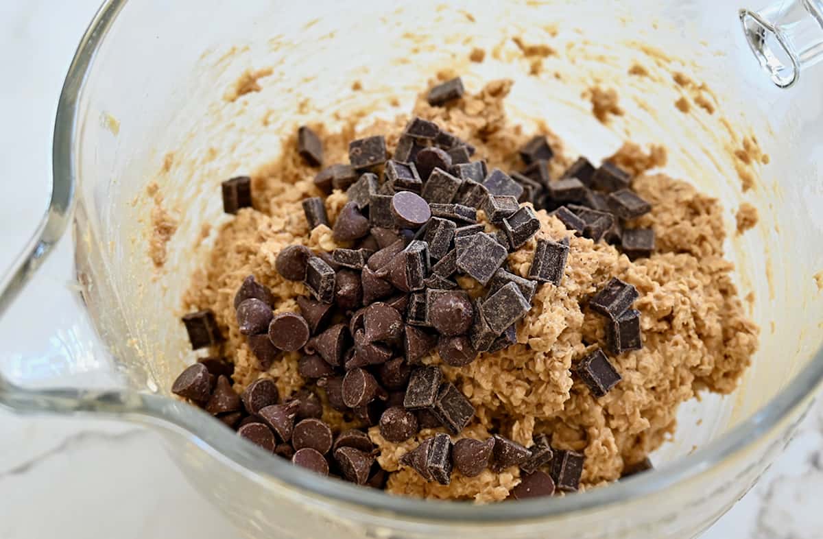 A large clear bowl containing oatmeal cookie dough topped with chocolate chips and chunks.