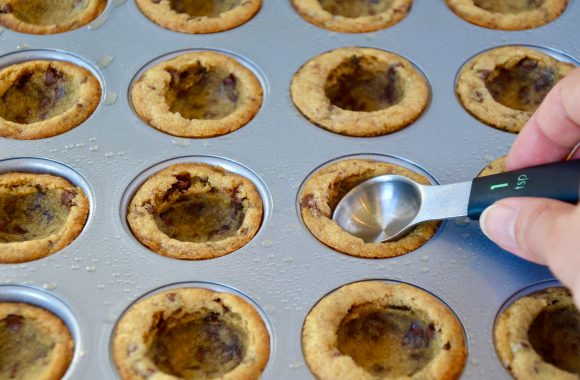 A hand holding a teaspoon measuring spoon making an indent in Chocolate Chip Cookie Cups