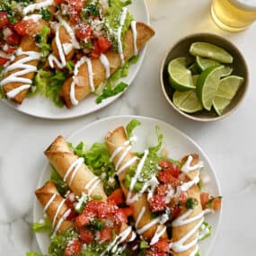 Two white plates with baked cheese taquitos topped with pico de gallo and guacamole piled atop a bed of lettuce next to a small bowl containing lime slices