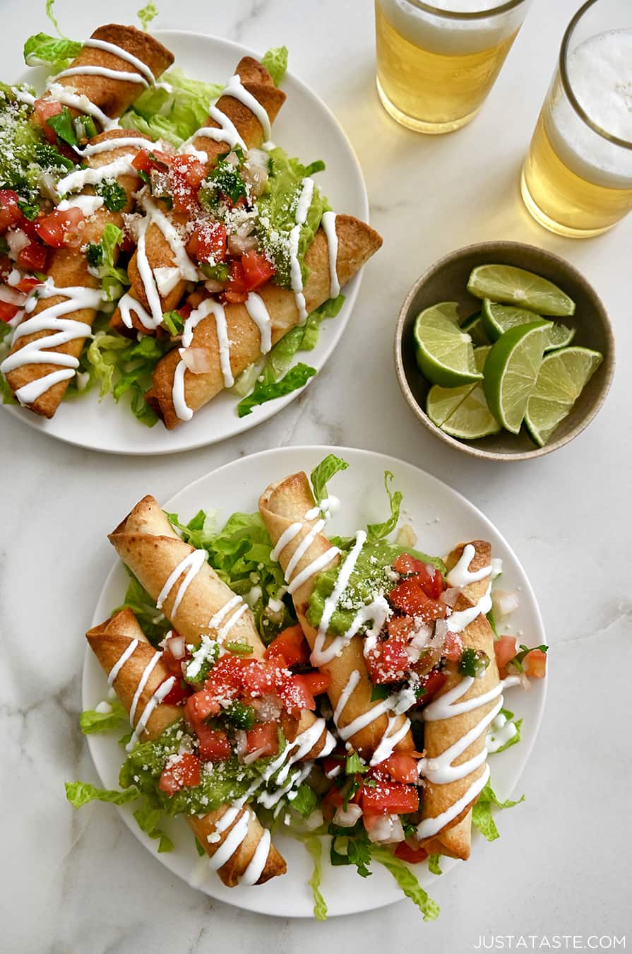 Two white plates with baked cheese taquitos topped with pico de gallo and guacamole piled atop a bed of lettuce next to a small bowl containing lime slices