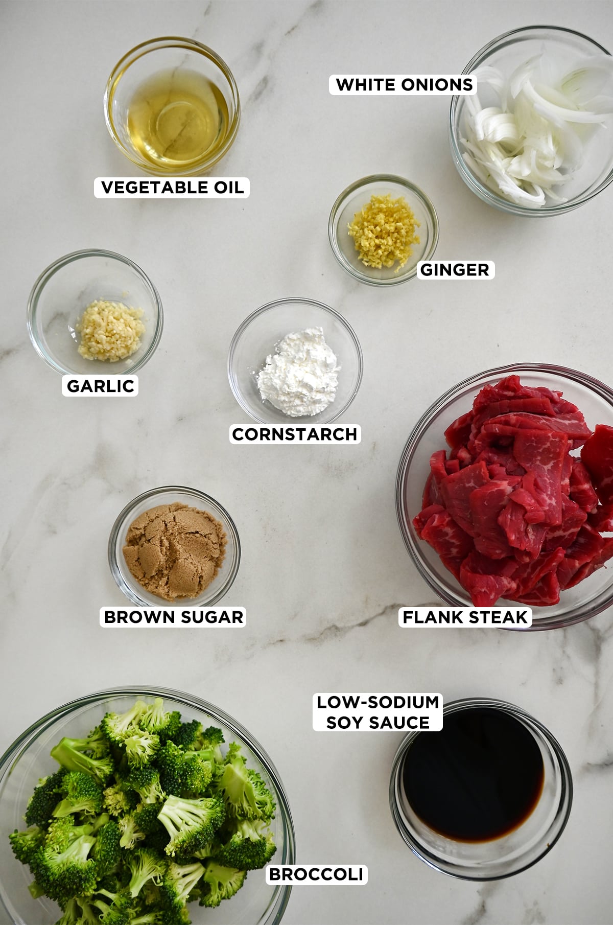 Various sizes of clear bowls containing sliced white onions, minced ginger, pieces of flank steak, low-sodium soy sauce, broccoli florets, brown sugar, cornstarch, minced garlic and vegetable oil.