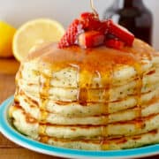 A tall stack of lemon poppyseed pancakes topped with strawberry slices that's all being drizzled with maple syrup.