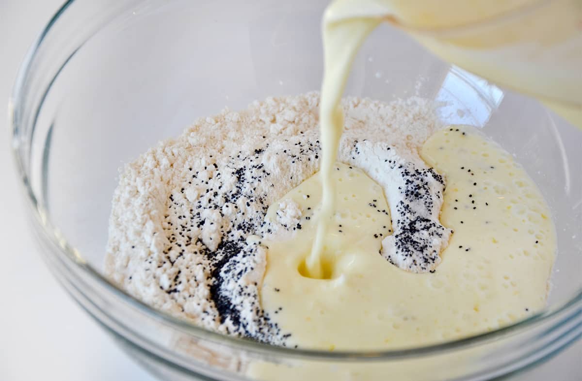 Wet ingredients being poured over flour and poppyseeds in a clear bowl.