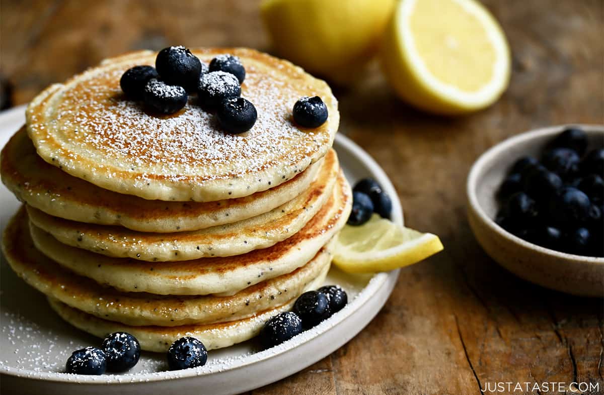 A tall stack of lemon poppyseed pancakes topped with blueberries and powdered sugar on a plate with blueberries and a lemon wedge.