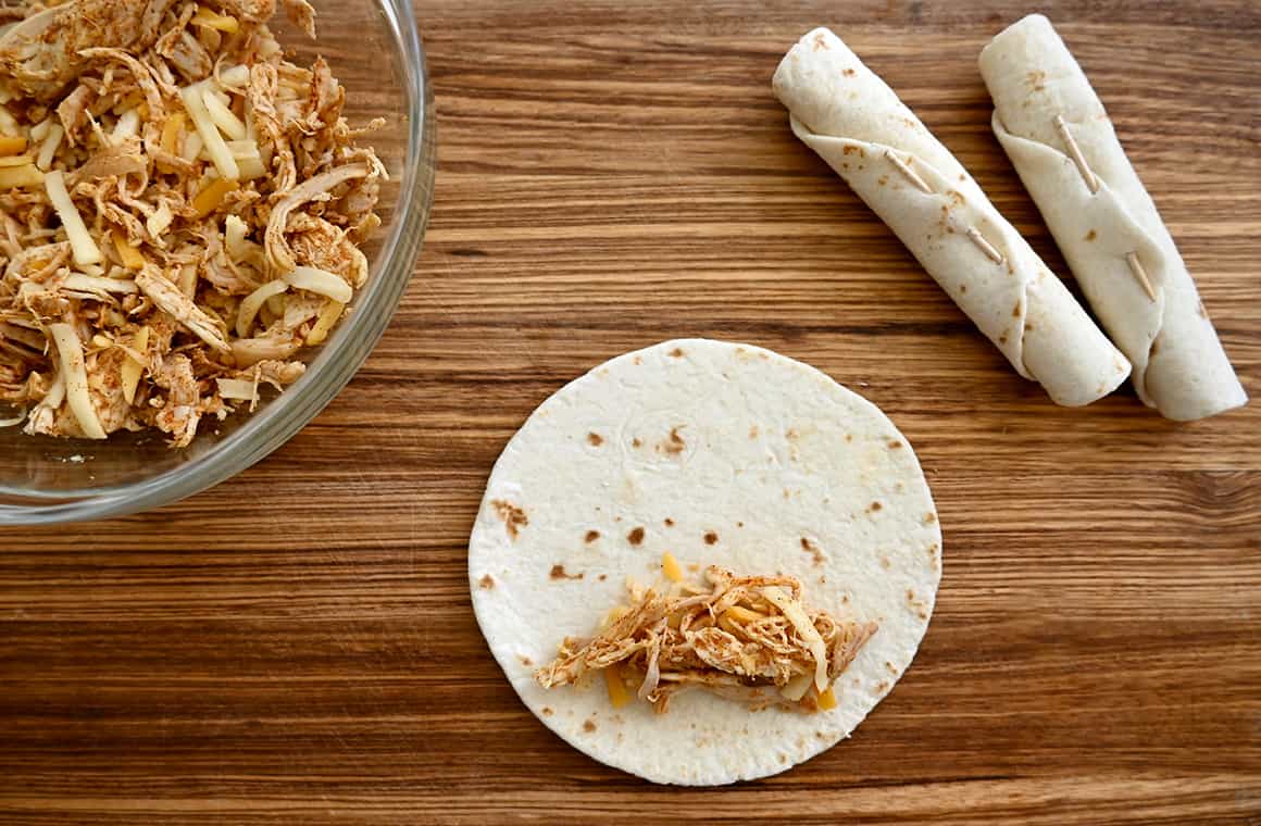 Chicken and cheese mixture atop a flour tortilla on a cutting board next to rolled-up tortillas
