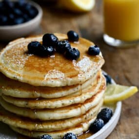 A tall stack of lemon poppyseed pancakes topped with fresh blueberries and drizzled with maple syrup.