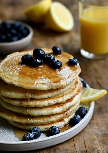 A tall stack of lemon poppyseed pancakes topped with fresh blueberries and drizzled with maple syrup.