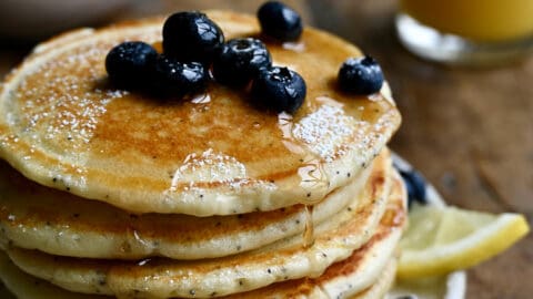 A tall stack of lemon poppyseed pancakes topped with fresh blueberries and drizzled with maple syrup.