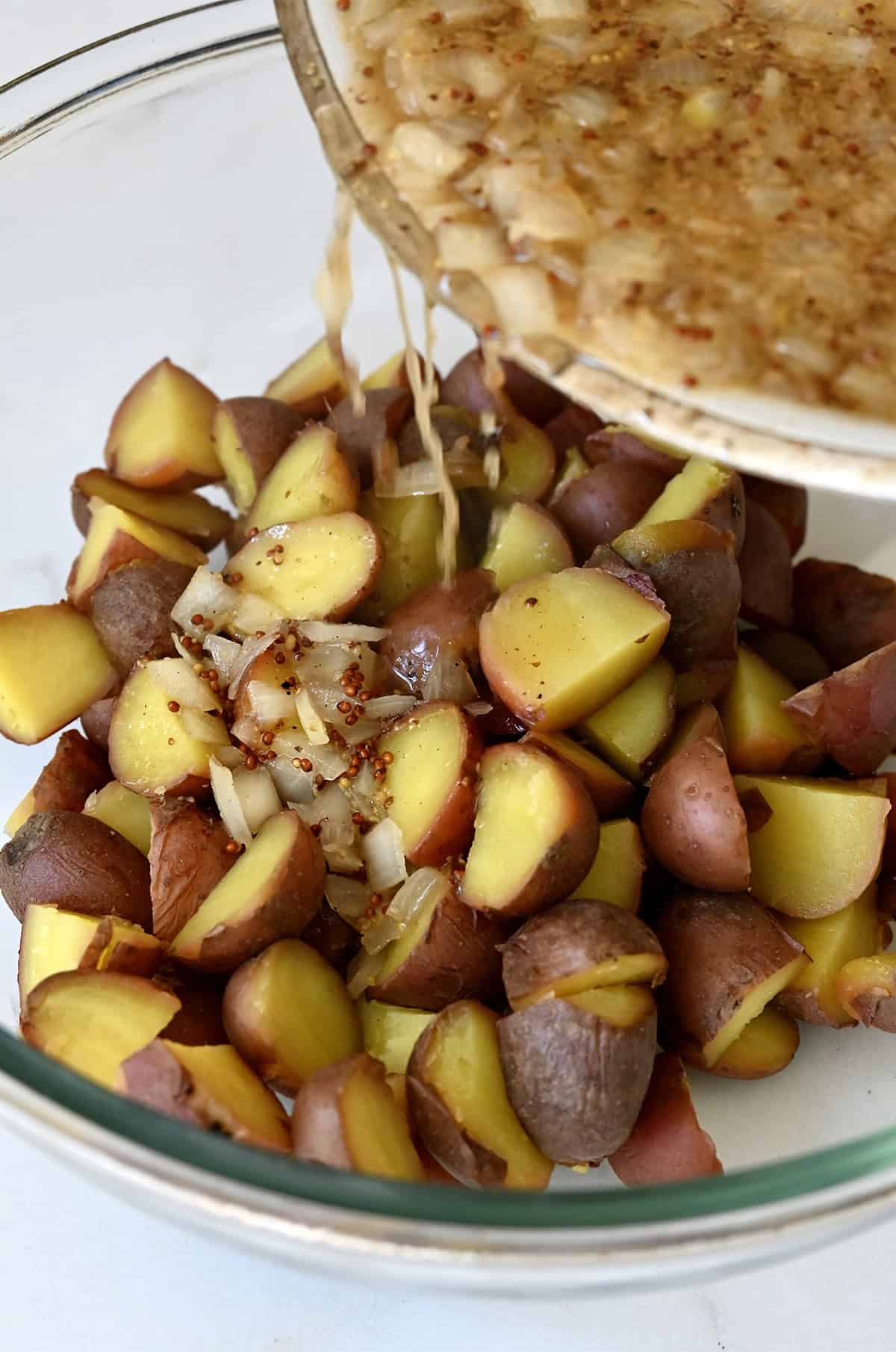 Pouring warm bacon dressing over boiled baby red potatoes in a large bowl.