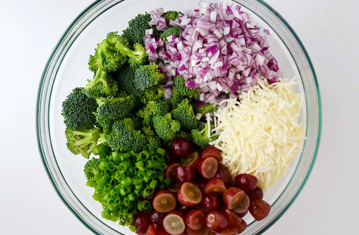 A top-down view of a clear bowl containing diced red onion, shredded mozzarella cheese, halved red grapes, chopped scallions and broccoli florets.