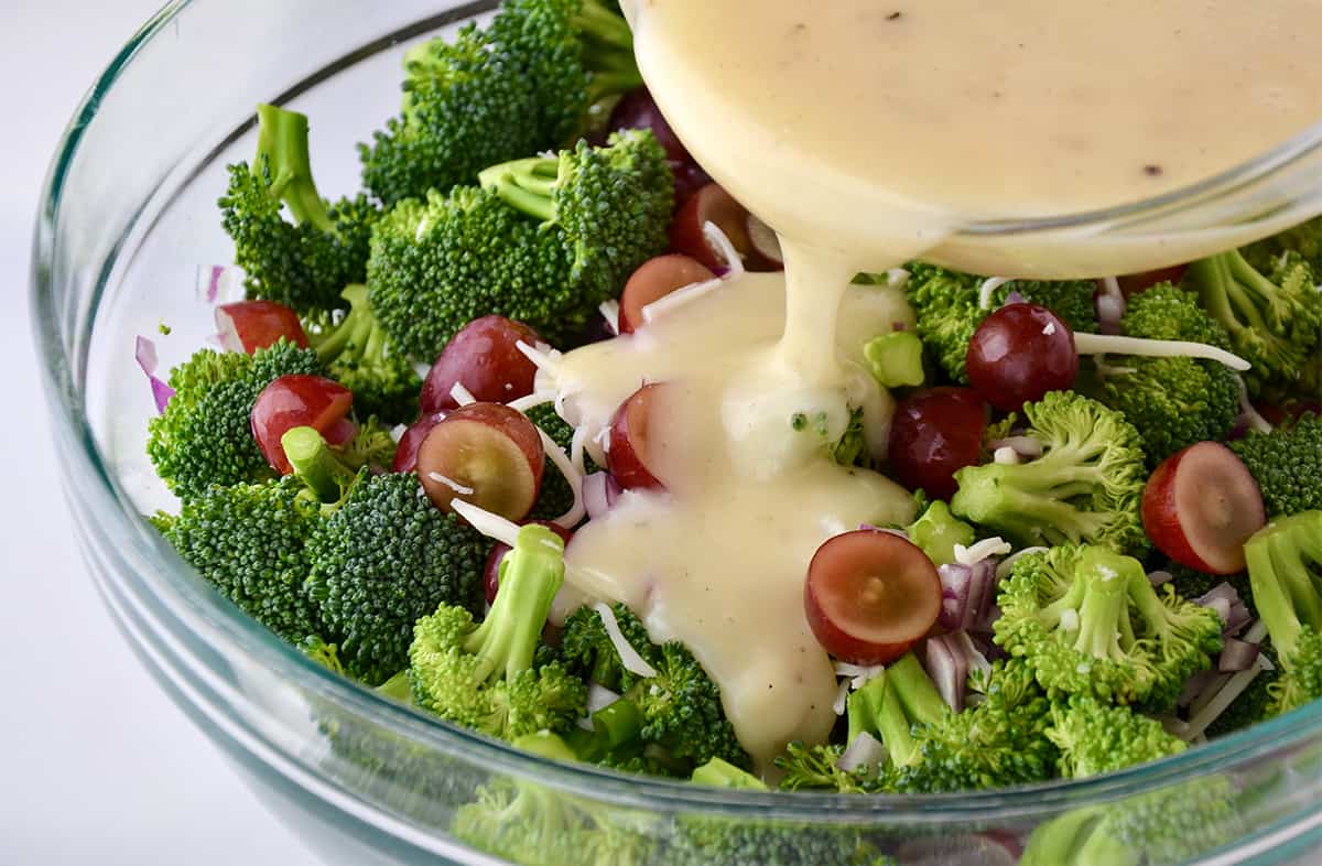 Creamy dressing being poured over broccoli florets, diced red onion and halved red grapes in a clear bowl.