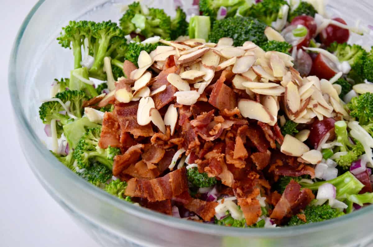 A large clear bowl containing broccoli salad topped with sliced almonds and crispy bacon pieces.