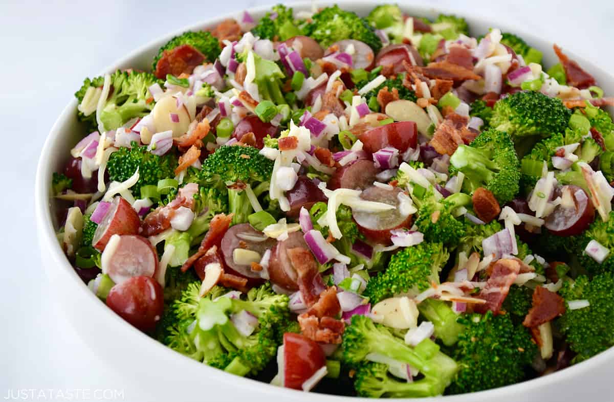 A large white serving bowl containing broccoli salad with creamy dressing, red grapes, diced onion, shredded mozzarella cheese and crispy bacon bits.