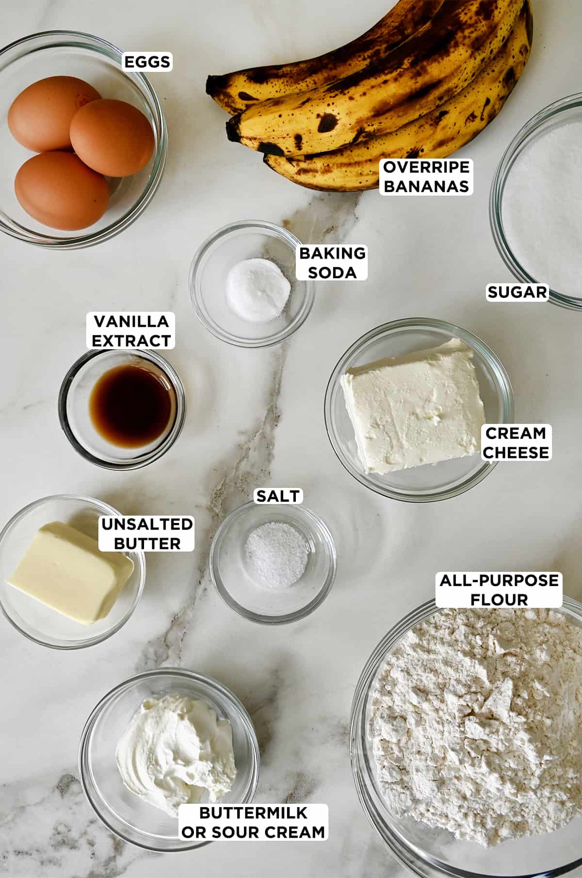 All of the ingredients needed to make cream cheese-swirled banana bread in glass bowls, including two eggs, three overripe bananas, granulated sugar, cream cheese, all-purpose flour, baking soda, salt, vanilla extract, unsalted butter and buttermilk.