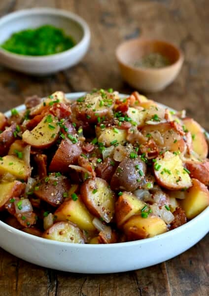 White serving bowl containing German potato salad with warm bacon dressing, bacon pieces and chopped fresh chives.
