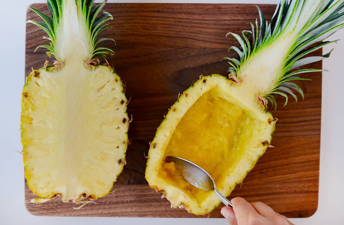 A hollowed out pineapple bowl next to it's other half on a wooden cutting board.