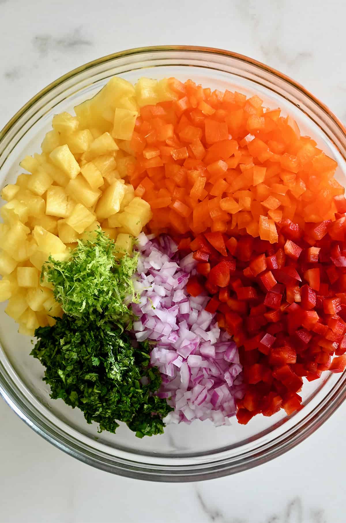 A large clear bowl containing chopped orange and red bell peppers, diced red onion, minced cilantro, lime zest and small pieces of pineapple.