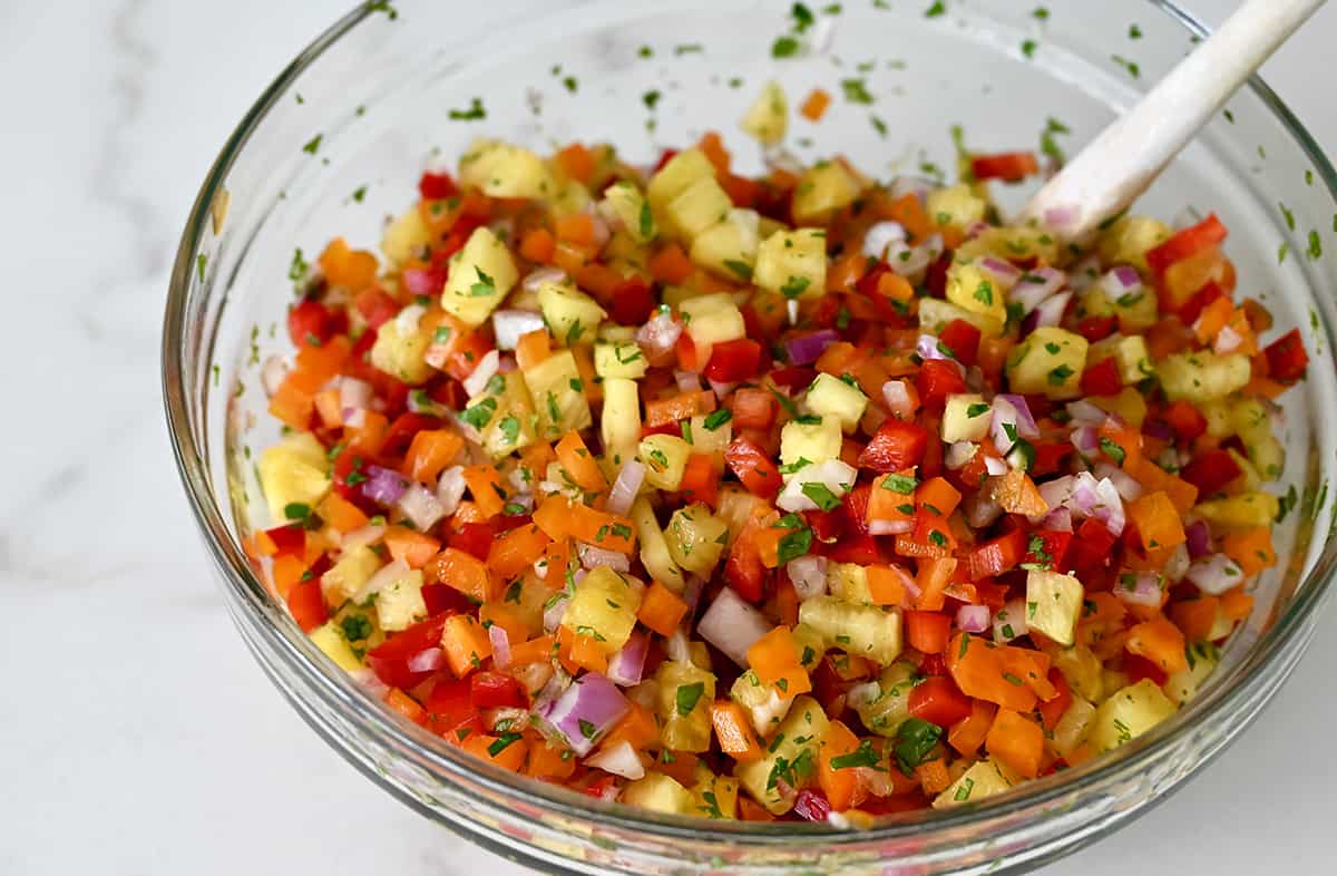 A clear bowl with a spoon containing pineapple salsa with bell peppers and fresh cilantro.