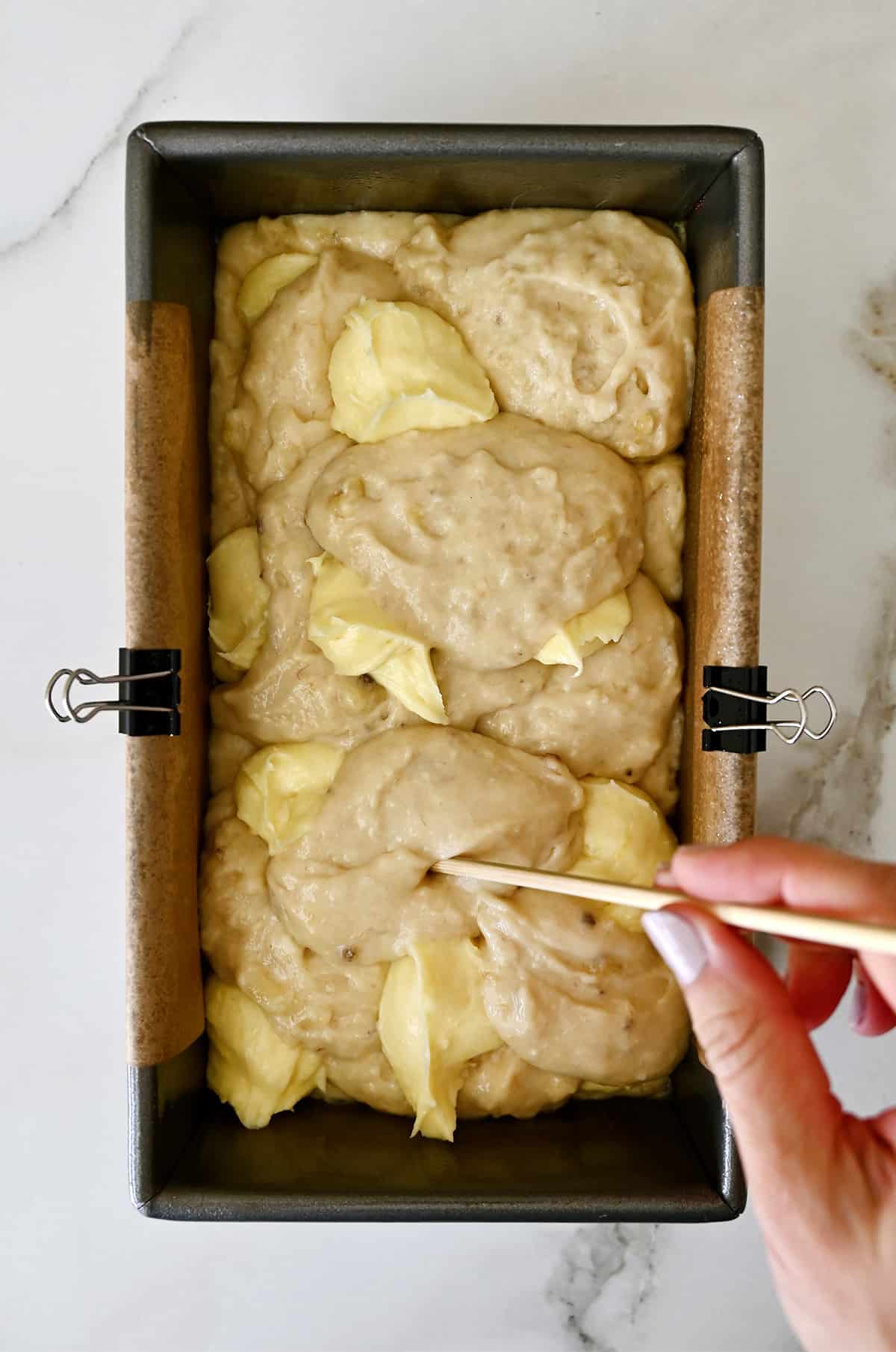 A hand holding a wooden skewer swirls cream cheese together with banana bread batter in a parchment paper-lined loaf pan.