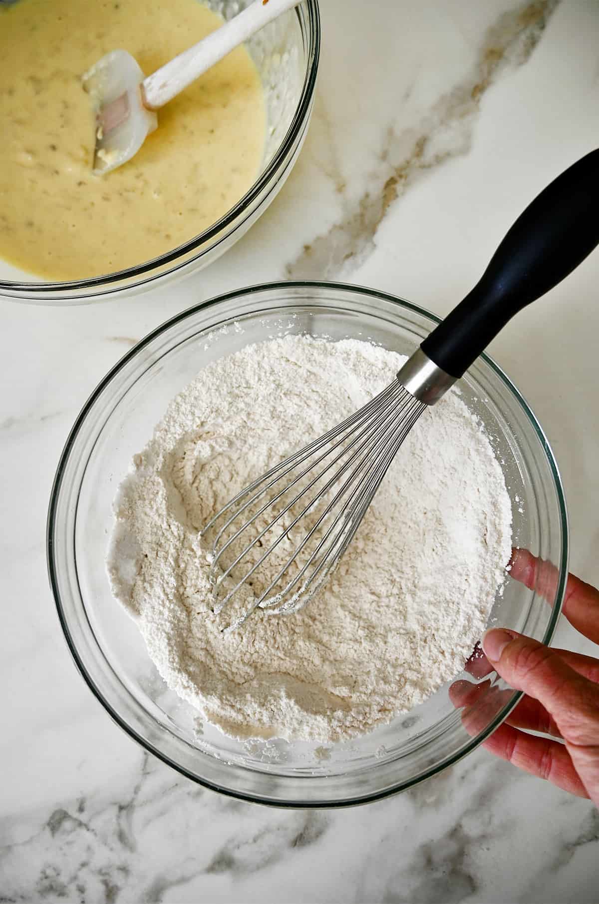 A glass bowl containing mashed bananas whisked together with eggs next to a glass bowl containing flour and a whisk. 