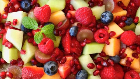 A large serving bowl containing fruit salad with diced cantaloupe and honeydew melon, blueberries, raspberries and pomegranate arils.