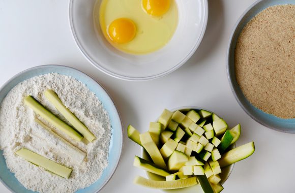 Zucchini fries in bowl with flour next to bowl with eggs and a bowl with breadcrumbs