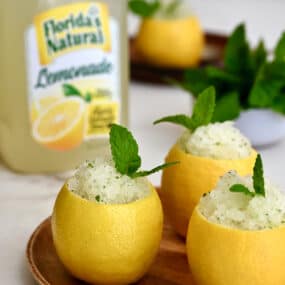 Three hollowed out lemons filled with lemon granita on a wood plate in front of a carton of Florida's Natural Lemonade.