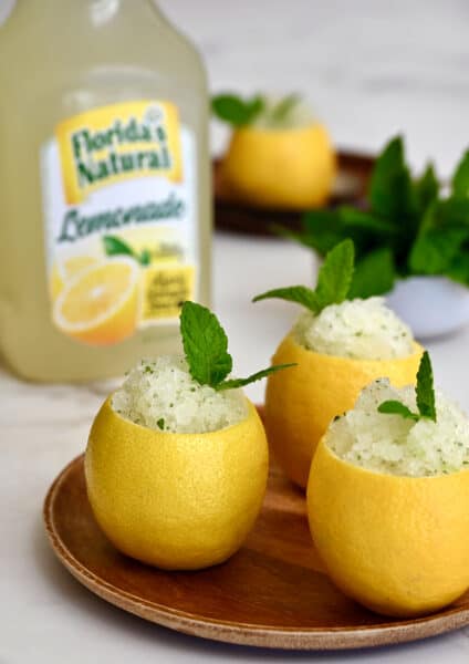 Three hollowed out lemons filled with lemon granita on a wood plate in front of a carton of Florida's Natural Lemonade.