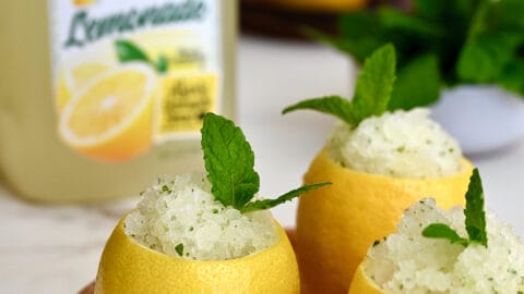 Three hollowed out lemons filled with lemon granita on a wood plate in front of a carton of Florida's Natural Lemonade.