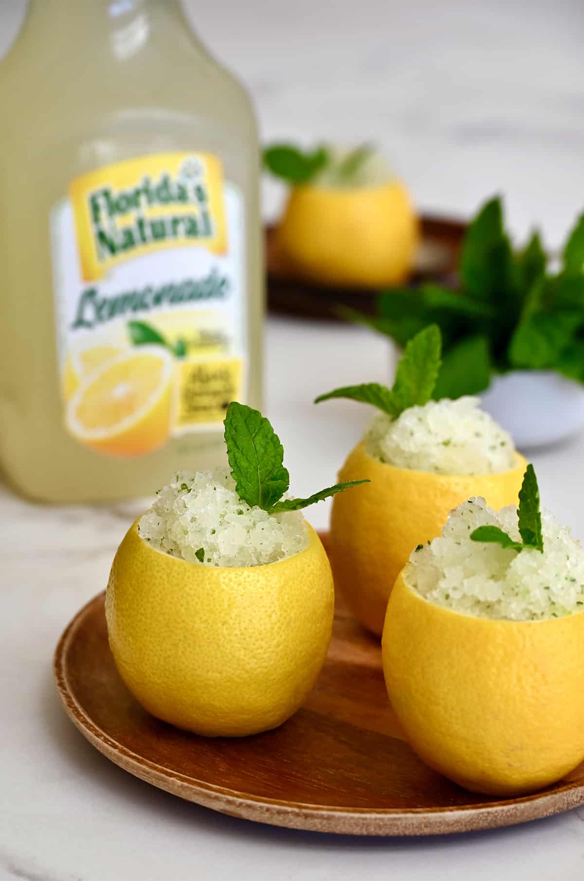 Three hollowed out lemons filled with lemon granita on a wood plate in front of a carton of Florida's Natural Lemonade.