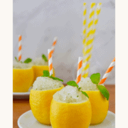 Three hollowed-out lemons filled with Easy Lemon Granita garnished with mint leaves and orange-and-white striped straws on a white plate.