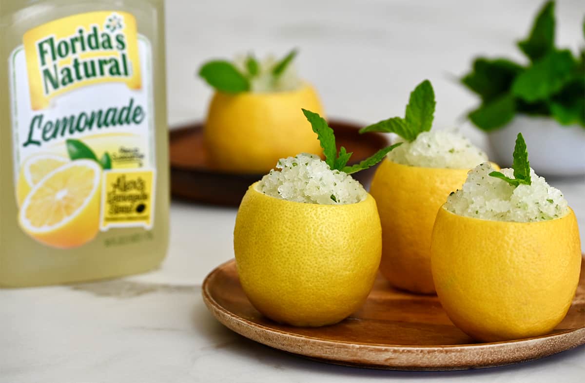 Lemon slushies in hollowed out lemons garnished with fresh mint on a plate next to a carton of Florida's Natural Lemonade.