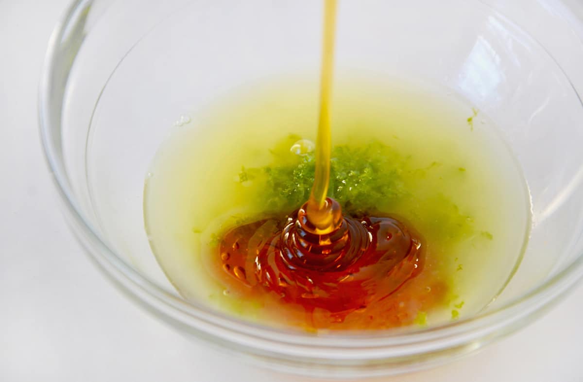 Honey being poured into a clear bowl that contains lime juice and lime zest.