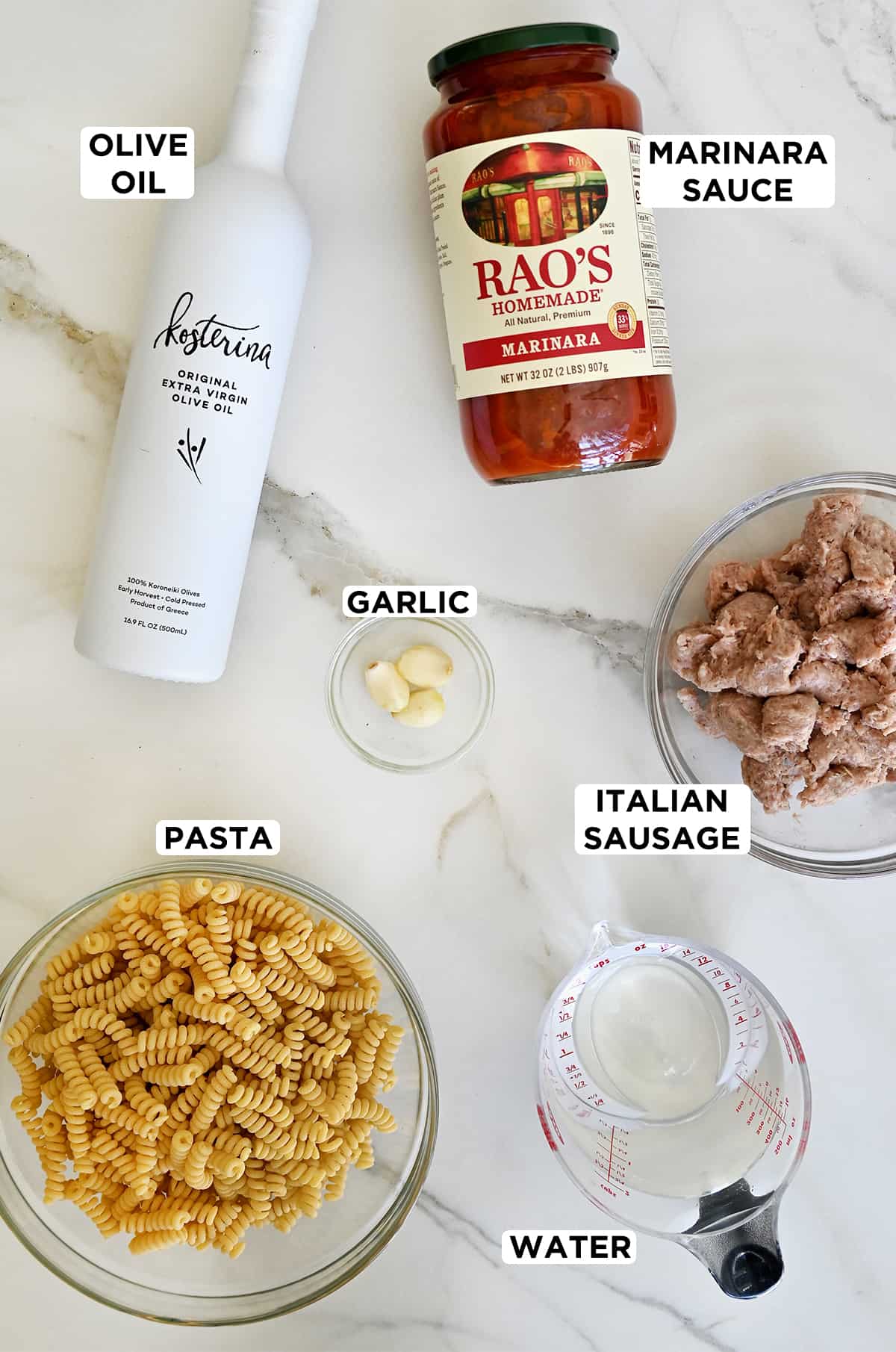 A bottle of olive oil next to a jar of marinara sauce, Italian sausage in a clear bowl, a measuring cup containing water, a clear bowl containing uncooked pasta and a small clear bowl containing minced garlic.