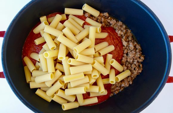 Uncooked noodles in pot with marinara sauce and cooked sausage