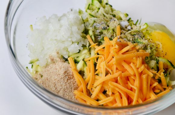 Shredded zucchini, diced onion, breadcrumbs, eggs, and shredded cheddar cheese in clear bowl