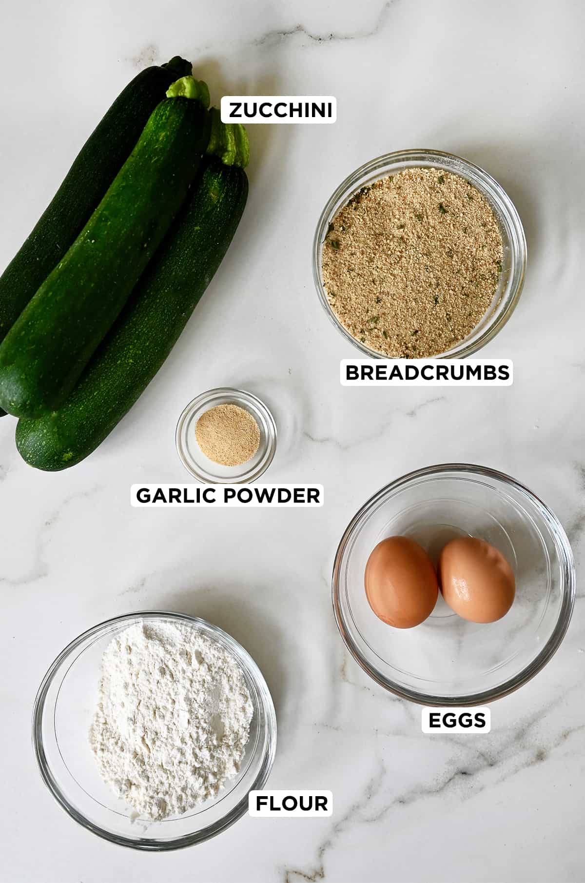 Three zucchini, a small bowl containing breadcrumbs, a bowl with two whole eggs, a bowl with flour and a small bowl containing garlic powder.