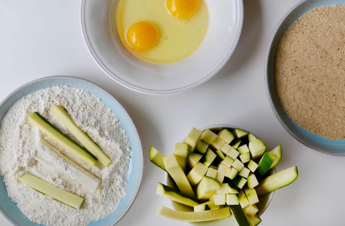 A bowl containing flour and zucchini sticks next to a bowl containing two eggs, a plate with breadcrumbs and a cup filled with zucchini sticks.